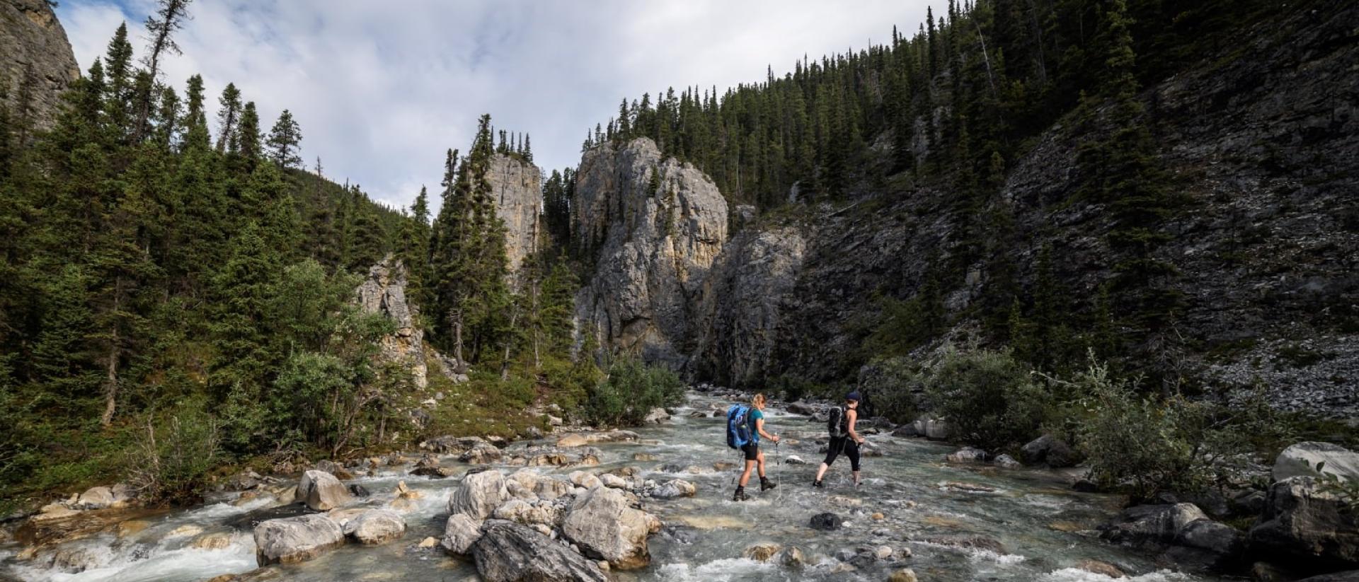 Two people hiking across shallow river with tree-covered rocky terrain in the background